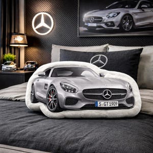 Può includere: Un cuscino a forma di auto sportiva Mercedes-Benz argentata poggia su un copriletto grigio. Il cuscino ha ruote nere con pinze freno rosse. Una foto incorniciata dell'auto è appesa al muro, insieme a un logo Mercedes-Benz.