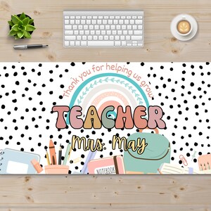 May include: White desk mat with black polka dots. It features a rainbow with the text "Thank you for helping us grow."  The word "TEACHER" is in colorful, bubble letters. The text "Mrs. May" is below the word "TEACHER".  There are school supplies on the bottom of the mat, including a pencil holder, a notebook, a backpack, and a binder.