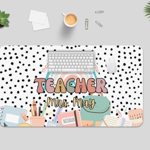 May include: A white desk mat with black polka dots and the text "Teacher Mrs. May". The mat features a rainbow, a backpack, pencils, and other school supplies.
