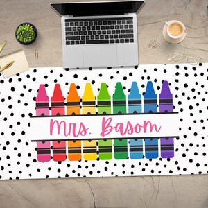 May include: A white desk mat with black polka dots and a row of rainbow colored crayons with the text "mrs. Basom" in pink.
