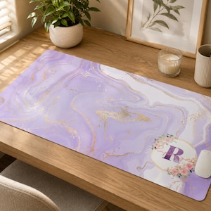 May include: A rectangular desk mat featuring a lavender and white marble pattern with gold veining accents. The bottom right corner displays a circular emblem containing the letter R, surrounded by a delicate floral wreath of pink and green.