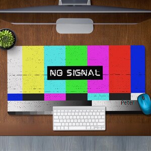 May include: A colorful computer mouse pad with a retro television static pattern and the text "NO SIGNAL" in a black rectangle. The mouse pad is on a wooden desk with a keyboard and a cup of coffee.