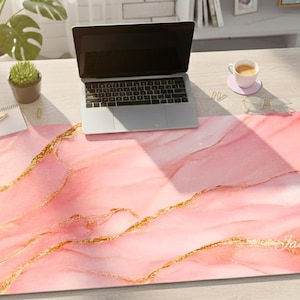 May include: A pink and gold desk mat with a marble design. The mat has a light pink background with gold veins and the name "Jasmine" in a script font. A laptop sits on the mat.
