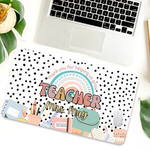 May include: A white laptop desk mat with black polka dots. The mat features a rainbow with the word "TEACHER" in colorful letters and the text "Thank you for helping us grow."  The text "Mrs. May" is written below the rainbow. The mat also includes a variety of school supplies, including a notebook, pencils, crayons, and a backpack.