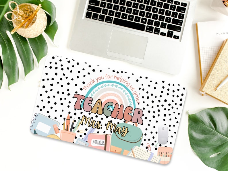 May include: A white laptop desk mat with black polka dots. The mat features a rainbow with the word "TEACHER" in colorful letters and the text "Thank you for helping us grow."  The text "Mrs. May" is written below the rainbow. The mat also includes a variety of school supplies, including a notebook, pencils, crayons, and a backpack.