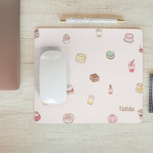 May include: A pink mouse pad with a variety of cute food illustrations, including cakes, pastries, and drinks. The mouse pad has the name "Natalia" printed on it.