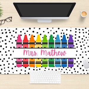 May include: A white desk mat with a black polka dot pattern. A row of colorful crayons is centered on the mat with the text "mrs. mathew" in pink lettering.