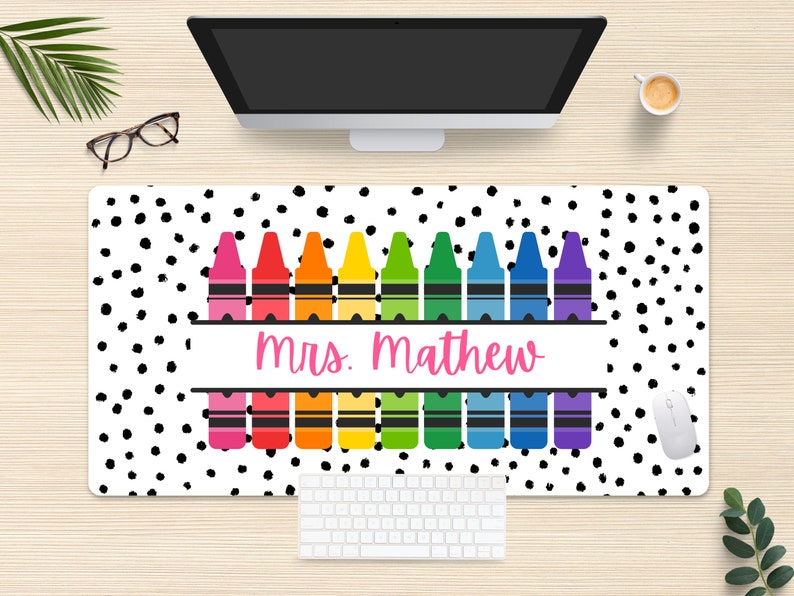 May include: A white desk mat with a black polka dot pattern. A row of colorful crayons is centered on the mat with the text "mrs. mathew" in pink lettering.