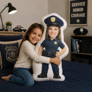 May include: A custom-printed pillow in the shape of a police officer, featuring a smiling face, a dark blue uniform, and a police hat with a gold badge. The pillow is being hugged by a young girl. The pillow is on a navy blue bed.