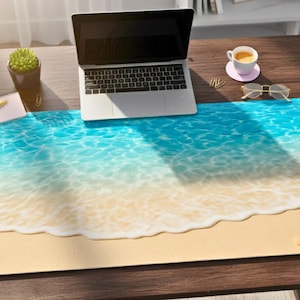 May include: A rectangular desk mat with a beach design, showing a blue ocean and sandy shore. A laptop, coffee cup, and stationery are on a wooden desk. The mat is approximately 76 cm long.