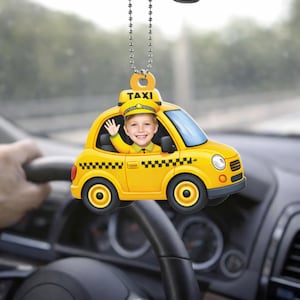 May include: A yellow taxi car-shaped car charm with a smiling child's face in the driver's seat. The charm has a black and white checkered pattern and the word "TAXI" on top. It hangs from a silver chain.