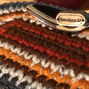 May include: A handmade, crocheted coaster in a mix of colors including gray, white, orange, and brown. A silver spoon rests on the coaster, with a small wooden tag that reads "Handmade". The coaster is likely used for a mug or bowl.