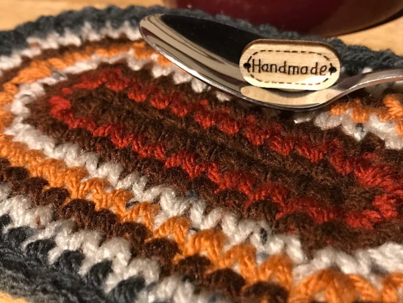 May include: A handmade, crocheted coaster in a mix of colors including gray, white, orange, and brown. A silver spoon rests on the coaster, with a small wooden tag that reads "Handmade". The coaster is likely used for a mug or bowl.