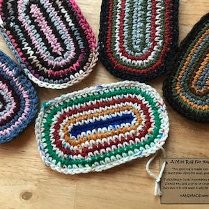 May include: A collection of handmade, oval-shaped mini rugs in various colors, including blue, pink, green, and brown. The rugs are crocheted with a textured design and are designed to hold a mug and a snack. A tag reads "A Mini Rug for your Mug."