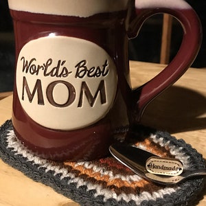 May include: A maroon ceramic mug with a cream-colored rim and handle, featuring the words "World's Best MOM" in a white oval. It rests on a handmade, multi-colored crochet coaster with a spoon that has a "Handmade" tag.
