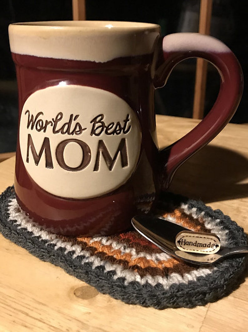 May include: A maroon ceramic mug with a cream-colored rim and handle, featuring the words "World's Best MOM" in a white oval. It rests on a handmade, multi-colored crochet coaster with a spoon that has a "Handmade" tag.