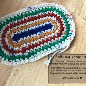May include: A handmade, oval-shaped mini rug for a mug. The rug features a colorful, crocheted design with a white border. The color scheme includes green, red, orange, blue, and white. The rug is designed to protect surfaces from spills.