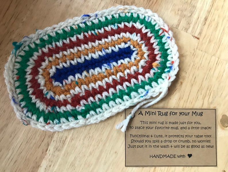 May include: A handmade, oval-shaped mini rug for a mug. The rug features a colorful, crocheted design with a white border. The color scheme includes green, red, orange, blue, and white. The rug is designed to protect surfaces from spills.