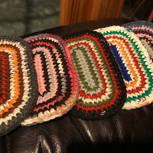 May include: A collection of five handmade, oval-shaped crochet rugs. Each rug features a unique color palette, including shades of brown, pink, green, blue, and white. The rugs are arranged side by side, showcasing their textured, handcrafted design.