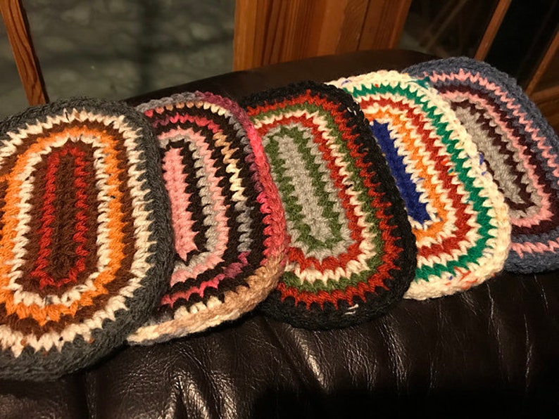 May include: A collection of five handmade, oval-shaped crochet rugs. Each rug features a unique color palette, including shades of brown, pink, green, blue, and white. The rugs are arranged side by side, showcasing their textured, handcrafted design.