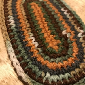 May include: An oval-shaped, handmade crochet item in earthy tones. The piece features concentric rings of brown, orange, green, and gray yarn, creating a textured surface. The edges are finished with a light green border.
