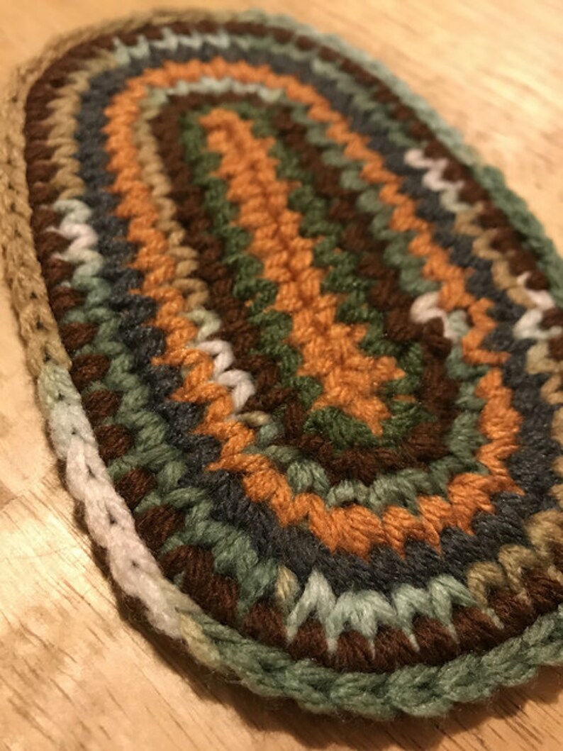 May include: An oval-shaped, handmade crochet item in earthy tones. The piece features concentric rings of brown, orange, green, and gray yarn, creating a textured surface. The edges are finished with a light green border.
