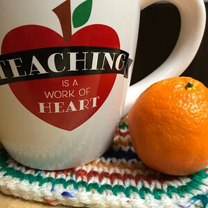 May include: A white ceramic mug with a red apple heart design and the text "TEACHING IS A WORK OF HEART." The mug sits on a handmade, colorful crochet coaster next to a bright orange tangerine.