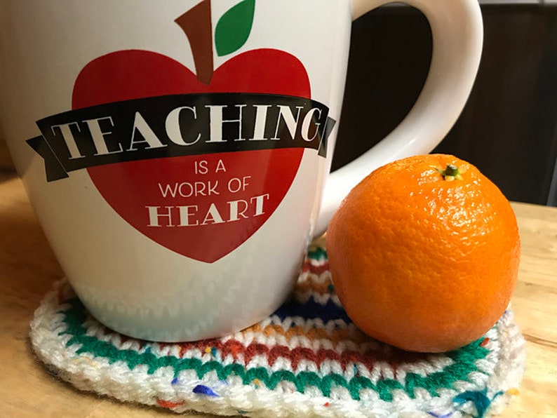May include: A white ceramic mug with a red apple heart design and the text "TEACHING IS A WORK OF HEART." The mug sits on a handmade, colorful crochet coaster next to a bright orange tangerine.