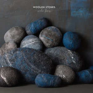 May include: A pile of blue and gray felt stones, arranged in a pyramid shape. The stones are made of wool and have a soft, textured surface. The text "WOOLEN STONES - woolen stories -" is visible at the top of the image.