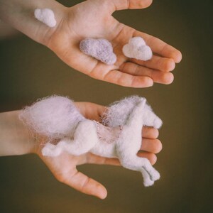 May include: A white felt unicorn with a pink felt saddle and mane, held in a person's hand. Three small felt clouds, two white and one purple, are also shown in the image. The text "WOOLEN STORIES - woolen stones -" is at the bottom of the image.