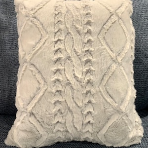 May include: A white faux fur throw pillow with a cable knit pattern. The pillow is square and has a soft, plush texture.