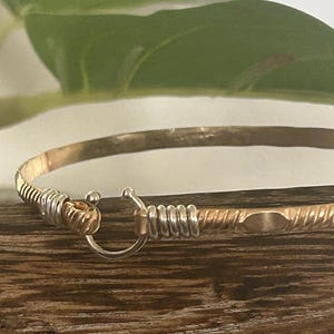 May include: A gold-toned bangle bracelet with silver accents. The bracelet features a hook and eye closure. The metal has a textured, twisted design. The bracelet is displayed on a wooden surface.