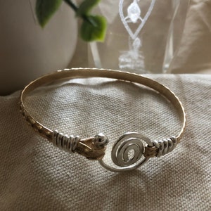 May include: A gold and silver-toned bangle bracelet. The bracelet features a textured gold band with silver wire detailing and a silver spiral clasp. The jewelry is displayed on a neutral-toned fabric.
