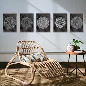 May include: Five black and white mandala prints with white outlines on a black background. The prints are arranged in a row on a white wall. A wicker lounge chair with a patterned cushion sits in front of the prints. A small black side table with a wooden top sits to the right of the chair. A pink mug and a potted plant are on the table.