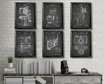 May include: Set of six black and white vintage camera patent prints framed in black frames. The prints feature different camera designs and are displayed on a white brick wall above a wooden shelf with books and a camera.