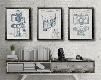 May include: Three vintage camera patent prints in black frames on a white brick wall. The prints are in blue and white and show different types of cameras. The prints are hanging above a white shelf with books and a vintage camera.