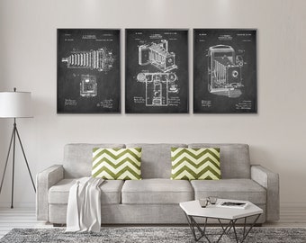 May include: Three black and white vintage camera patent prints with a gray background. The prints are framed and hung on a white wall above a gray couch with green pillows.