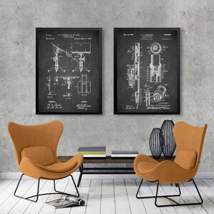 May include: Two black framed vintage patent prints on a gray wall. The left print shows a camera stand with the text "U.S. KIDER & W. L. LOWE" and "Patented May 17, 1917". The right print shows a camera lens with the text "Dec. 18, 1918" and "J.K.L. MILLER".