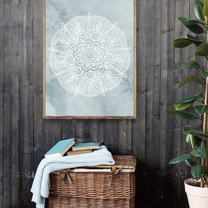 May include: A framed print of a white mandala design on a blue watercolor background. The mandala is intricate and detailed, with a floral pattern. The print is hanging on a dark gray wood wall.