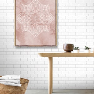 May include: A framed print with a white mandala design on a pink watercolor background. The print is hanging on a white subway tile wall above a wooden table with a brown ceramic pot and two small potted plants.