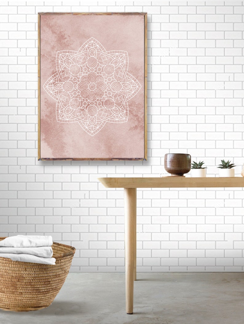 May include: A framed print with a white mandala design on a pink watercolor background. The print is hanging on a white subway tile wall above a wooden table with a brown ceramic pot and two small potted plants.