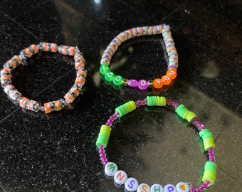 May include: Three colorful beaded bracelets. The first bracelet is orange and black with white beads. The second bracelet is green, purple, and orange with white beads that spell out "YOU". The third bracelet is green, purple, and yellow with white beads that spell out "MSHP" with a heart bead.