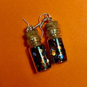Peut inclure: Une paire de boucles d'oreilles avec de petites bouteilles en verre remplies de paillettes colorées. Les bouteilles ont des bouchons en liège et des crochets argentés.