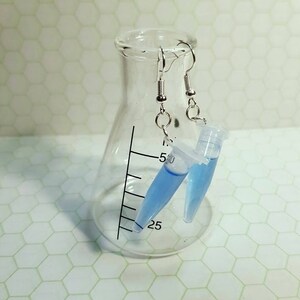 May include: A pair of silver earrings with blue liquid filled test tubes hanging from them. The earrings are displayed on a clear glass beaker.
