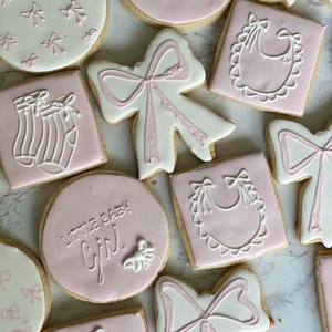 May include: Assortment of baby shower cookies with pink and white icing. The cookies feature designs of bows, baby bibs, and baby socks. Some cookies are round with the text "Little Baby Girl".