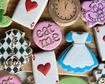 May include: Assorted decorated sugar cookies on a wooden surface. Designs include playing cards with hearts, a pocket watch, a dress, a Cheshire Cat face, a key, and a Mad Hatter hat. Some cookies have the text "eat me".