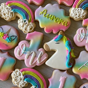 May include: A collection of decorated sugar cookies featuring a rainbow, unicorn, clouds, and the number one. The cookies are decorated with colorful frosting and sprinkles. The cookies are perfect for a first birthday party.