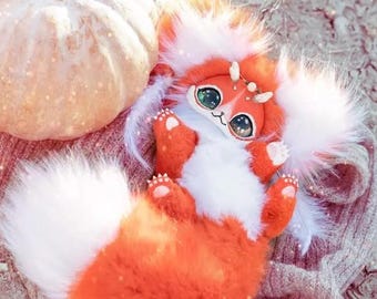 May include: A plush orange and white fantasy creature with large green eyes and a fluffy tail. The creature is lying on a textured surface next to a light-colored pumpkin. The creature has small horns and a sweet expression.