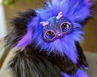 May include: A handmade fantasy creature with a plush, furry body in shades of black, purple, and blue. The creature has large, expressive eyes and a detailed face with small horns. The overall design is whimsical and detailed, with a focus on texture and color.