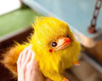 May include: A handmade plush creature with bright yellow fur, a reddish-orange beak, and large, dark eyes. The plush has a fluffy tail and small, detailed feet. The creature is held in a hand, showcasing its size and texture.